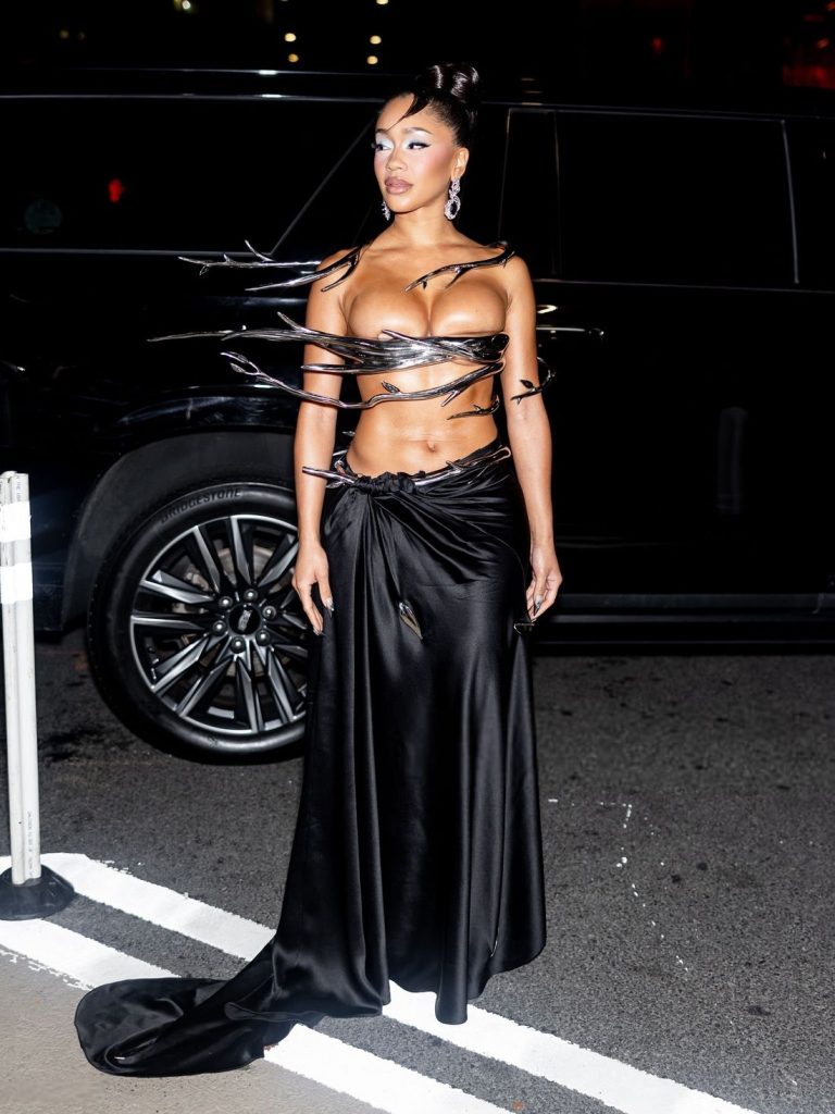 Woman in a high-fashion outfit with a metallic top featuring sharp, spiked extensions and a long, black satin skirt, standing outdoors at night in front of a black vehicle.