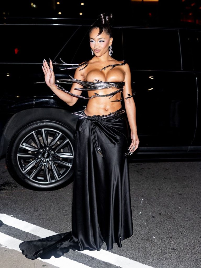A woman poses on the street at night wearing a striking black skirt and a sculptural silver and black top with metallic bands. She waves, and a black SUV is in the background.