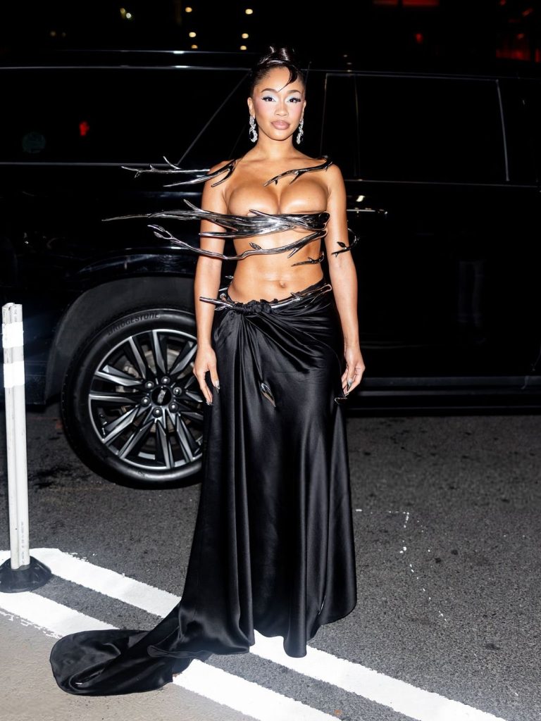 A woman stands outdoors at night wearing a striking black outfit with metallic elements resembling feathers, a long flowing skirt, and dramatic earrings. A dark vehicle is parked behind her.