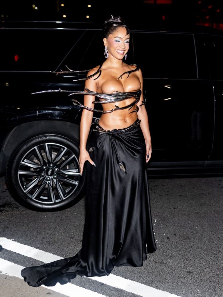 A woman in a unique black gown with a sculptural, cutout top and a draped skirt poses on a city street at night, smiling, with a black SUV in the background.