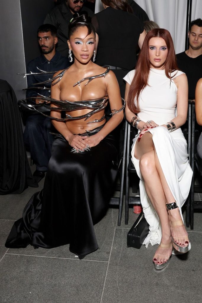 Two women sit side by side at a fashion event. One wears a dramatic black dress with metallic accents, exposing her torso. The other wears a sleek white dress with a high slit. Both have styled hair and wear high heels.
