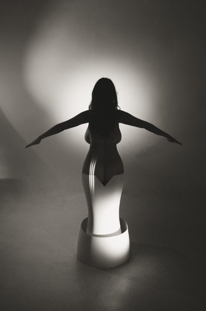 A silhouette of a person with arms outstretched stands inside a sculptural, vase-like structure. Light and shadow create dramatic contrast, highlighting the contours of the figure and the geometric shape.
