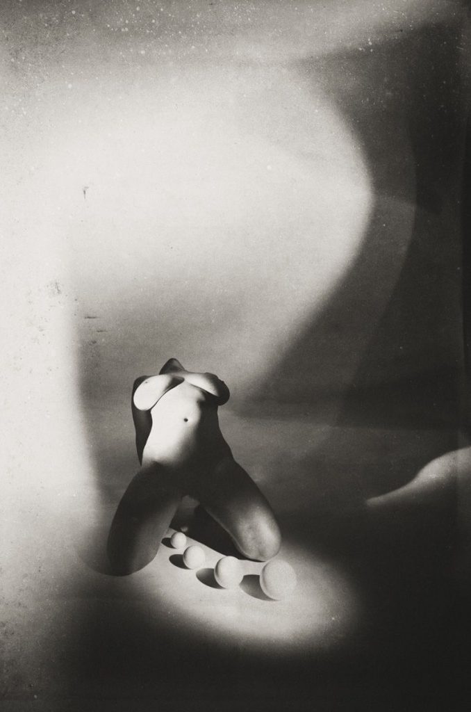 Black and white photo of a nude figure kneeling on the ground, head tilted back, with spherical objects placed in front. Abstract shadows and lighting create a surreal and dramatic atmosphere.