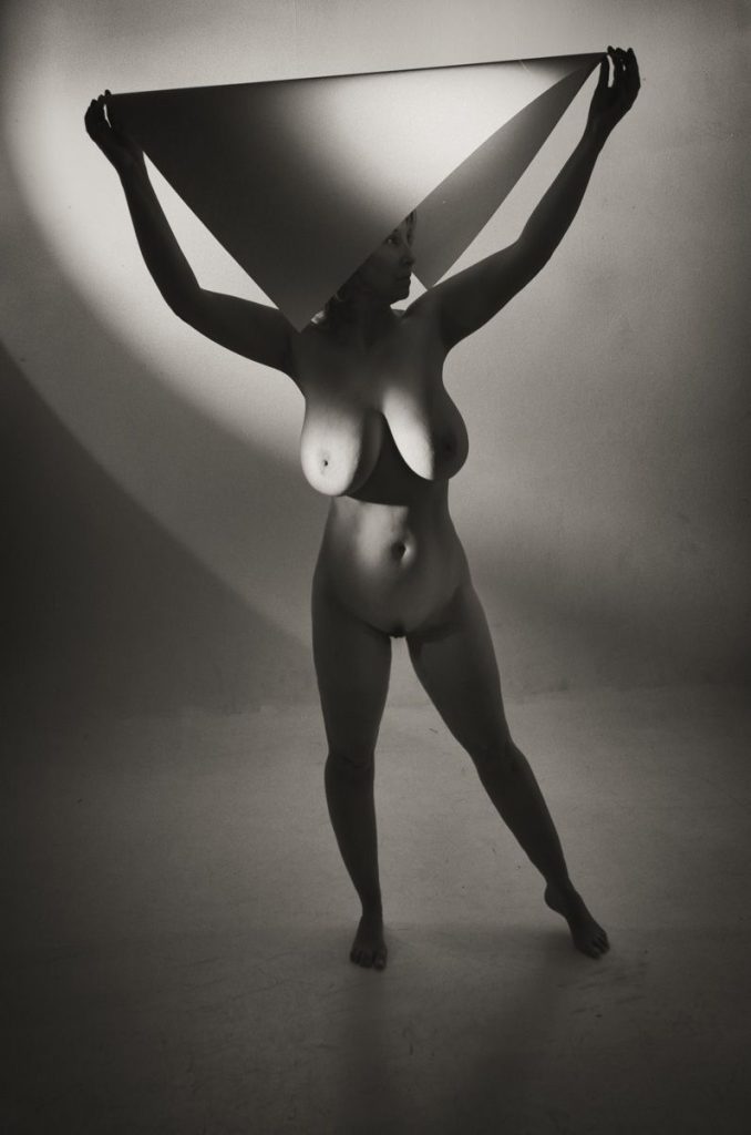 A nude person stands holding a large geometric cone above their head, casting dramatic shadows on the wall in a black-and-white, artistic photograph.