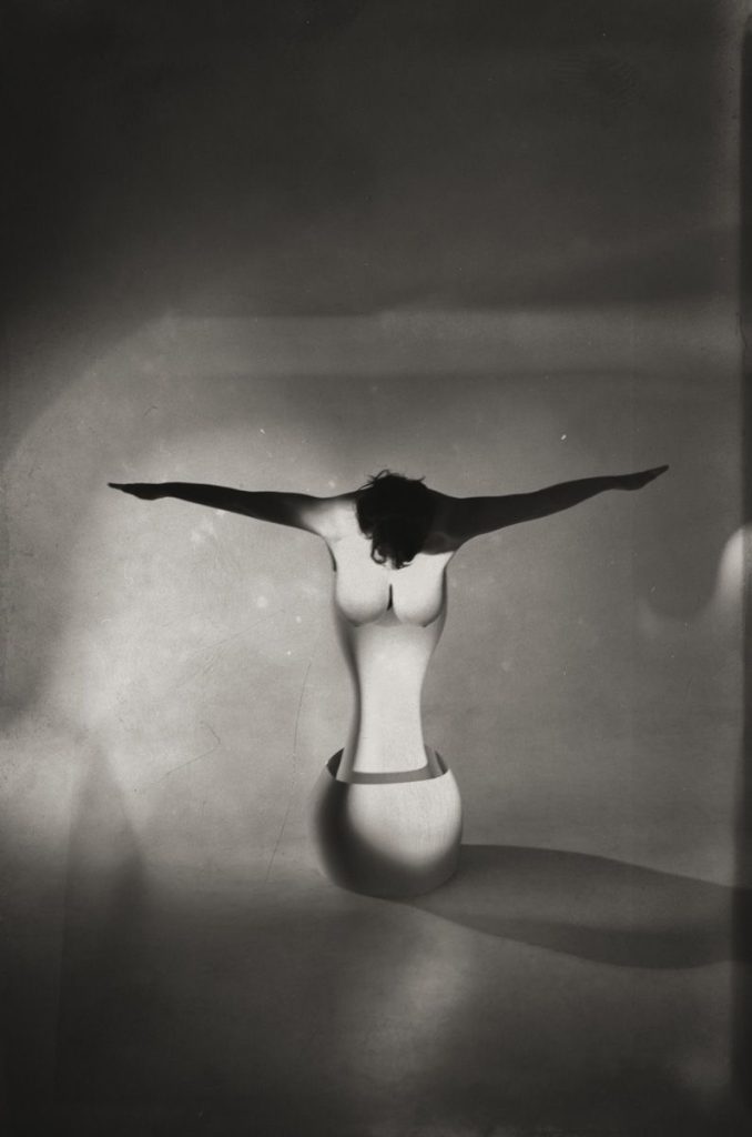 A womans torso emerges from a sculpted, headless mannequin form. Her arms are outstretched horizontally, casting dramatic shadows on a minimalist, dimly lit background.