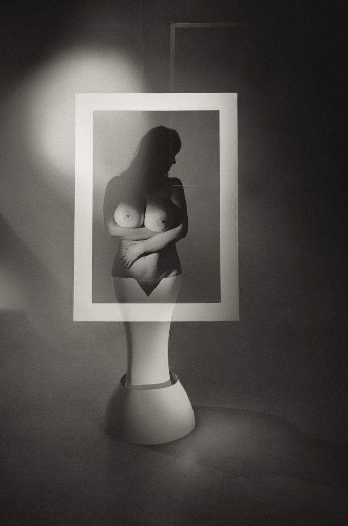 A grayscale photo features a nude figure with arms crossed, partially obscured by a rectangular frame, creating a layered, surreal effect with dramatic lighting and shadows.