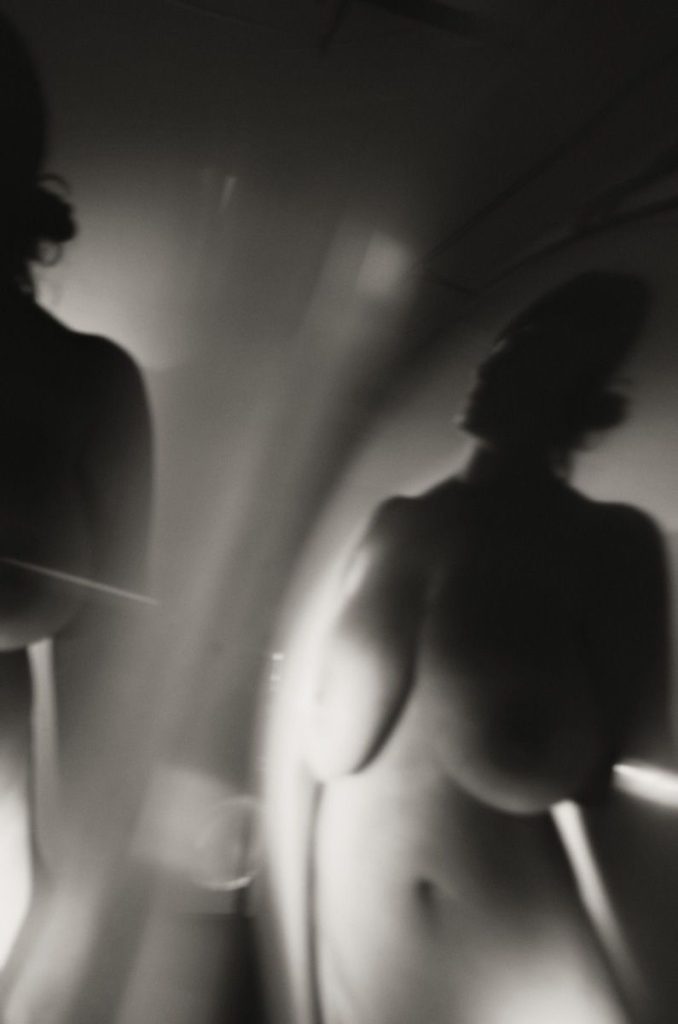 A blurred, abstract black and white image shows the artistic nude silhouettes of two people reflecting on a curved surface, creating a distorted and soft visual effect.