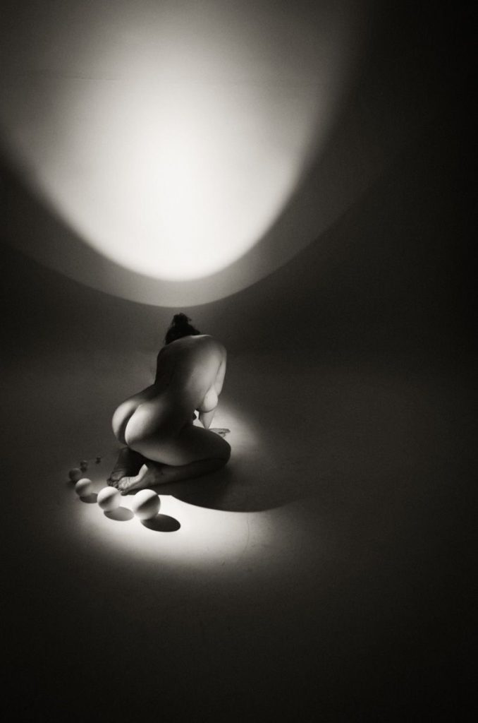A nude person kneels on the floor under a spotlight, facing away from the camera. Smooth stones are placed in a curved line nearby. The scene is in black and white with dramatic lighting and shadows.