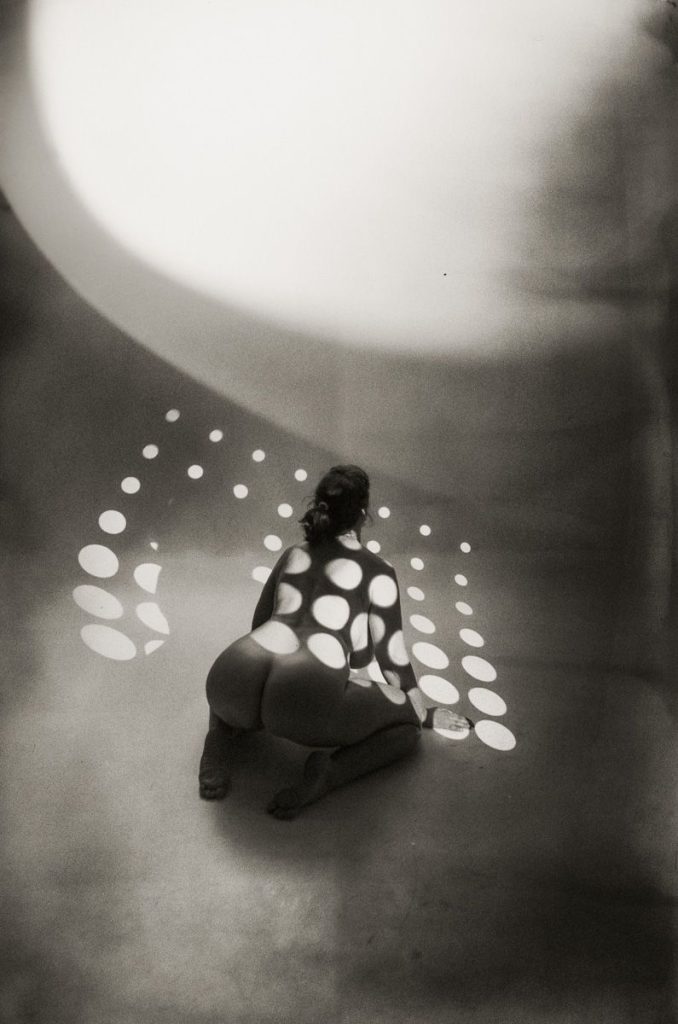 A person kneels on the floor under overhead, circular light patterns that cast dots on their body and the ground, creating an abstract, artistic scene in black and white.