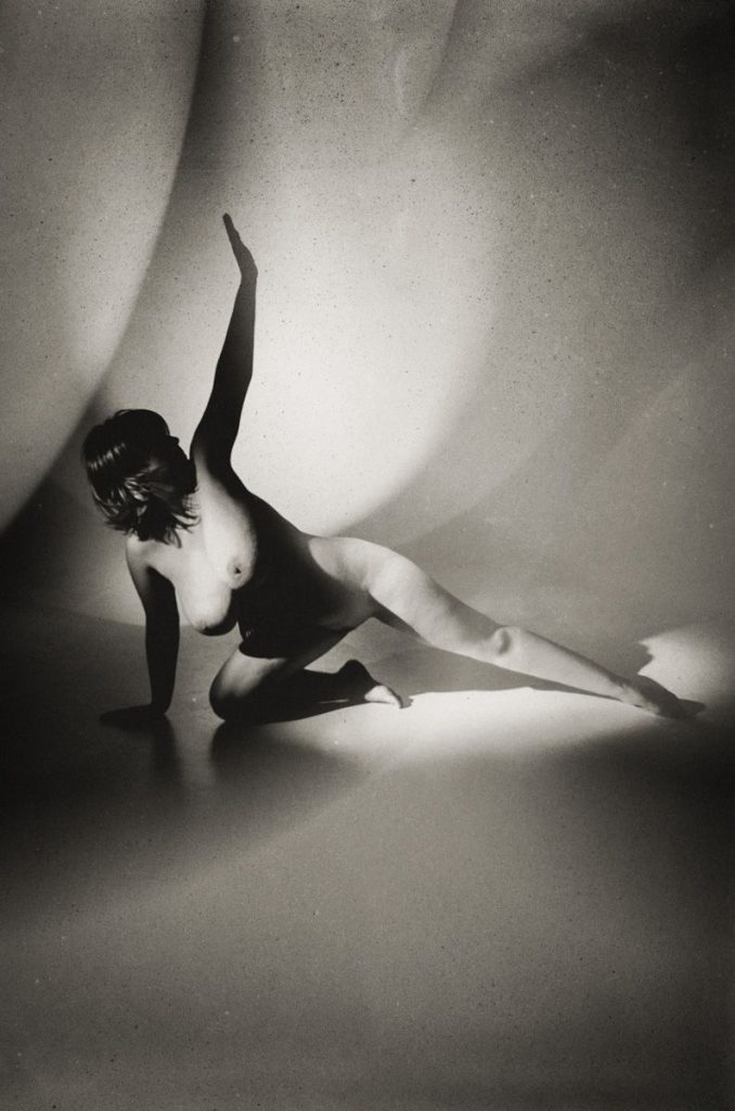 A nude person poses on the floor with one arm raised and one leg extended, dramatically lit in black and white with abstract shadows in the background.