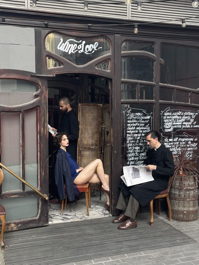 Three people outside a vintage-style café; one woman in a blue blazer sits on a chair, a man in black stands reading, and another man in a coat sits reading a newspaper. Handwritten menus decorate the entrance.