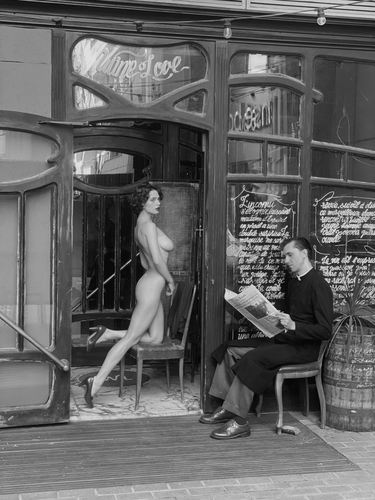 A nude woman poses on a chair at the entrance of a vintage café while a man in dark clothing sits outside reading a newspaper. The scene is in black and white.