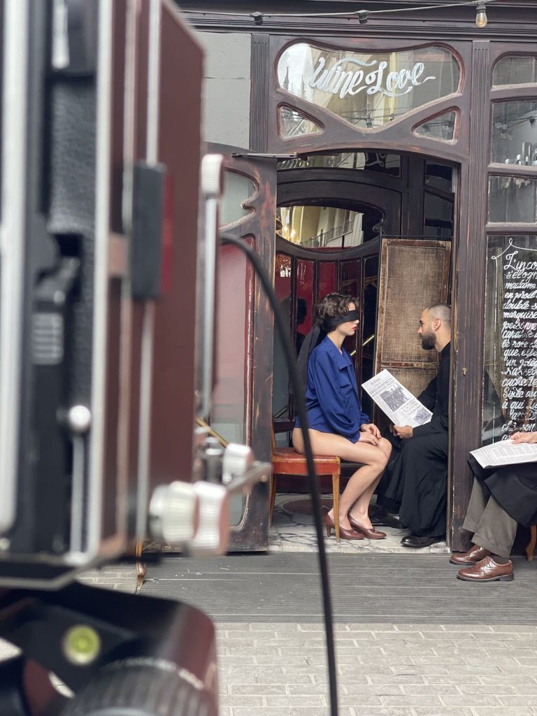 A man and a woman sit facing each other inside a café, holding menus. A large camera is visible in the foreground, and another person sits to the right, partially out of frame.