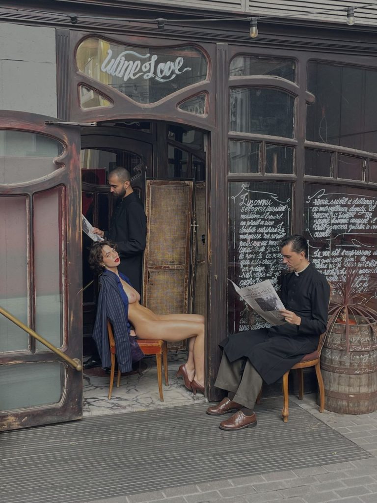 A half-naked woman in a blazer and heels poses seated in front of a cafe, while two men in dark clothing read newspapers nearby. The cafe has ornate glass doors and handwritten menus on the windows.