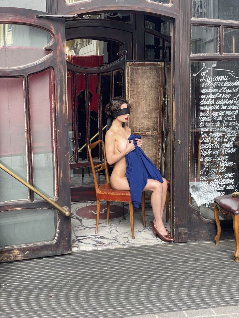A nude woman sits nude on a chair near an open doorway, holding a blue cloth to their chest. Her eyes are covered with a blingfold. The setting appears to be an ornate, vintage-style exterior.