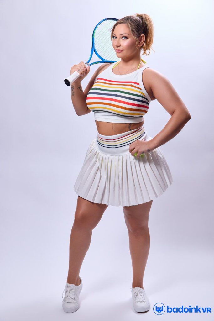 Woman with blonde hair in a ponytail standing in a white studio, wearing a rainbow-striped crop top and white pleated tennis skirt, holding a blue tennis racket over her shoulder.
