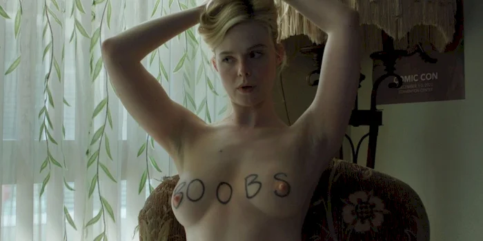 Elle Fanning topless with BOOBS written on her breasts. She twists and holds her hair up with both hands.