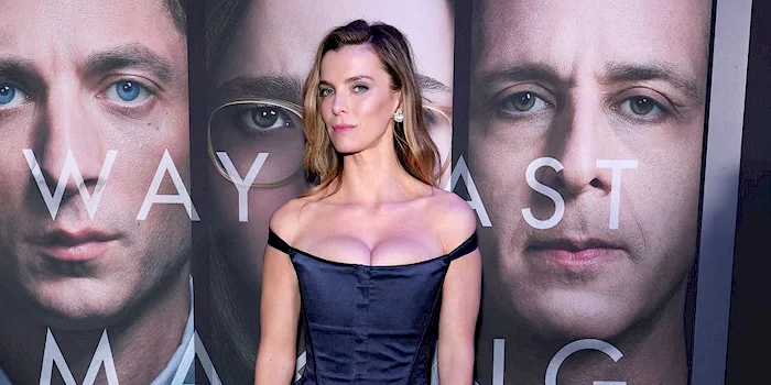 Betty Gilpin stands in front of a movie poster for Were Way Past Making Friends, wearing a dark corset-style dress.