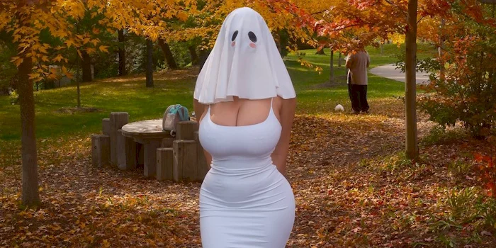 A woman in a ghost costume consisting of a white sheet with a ghost face worn over a white dress, standing in a park with autumn leaves.