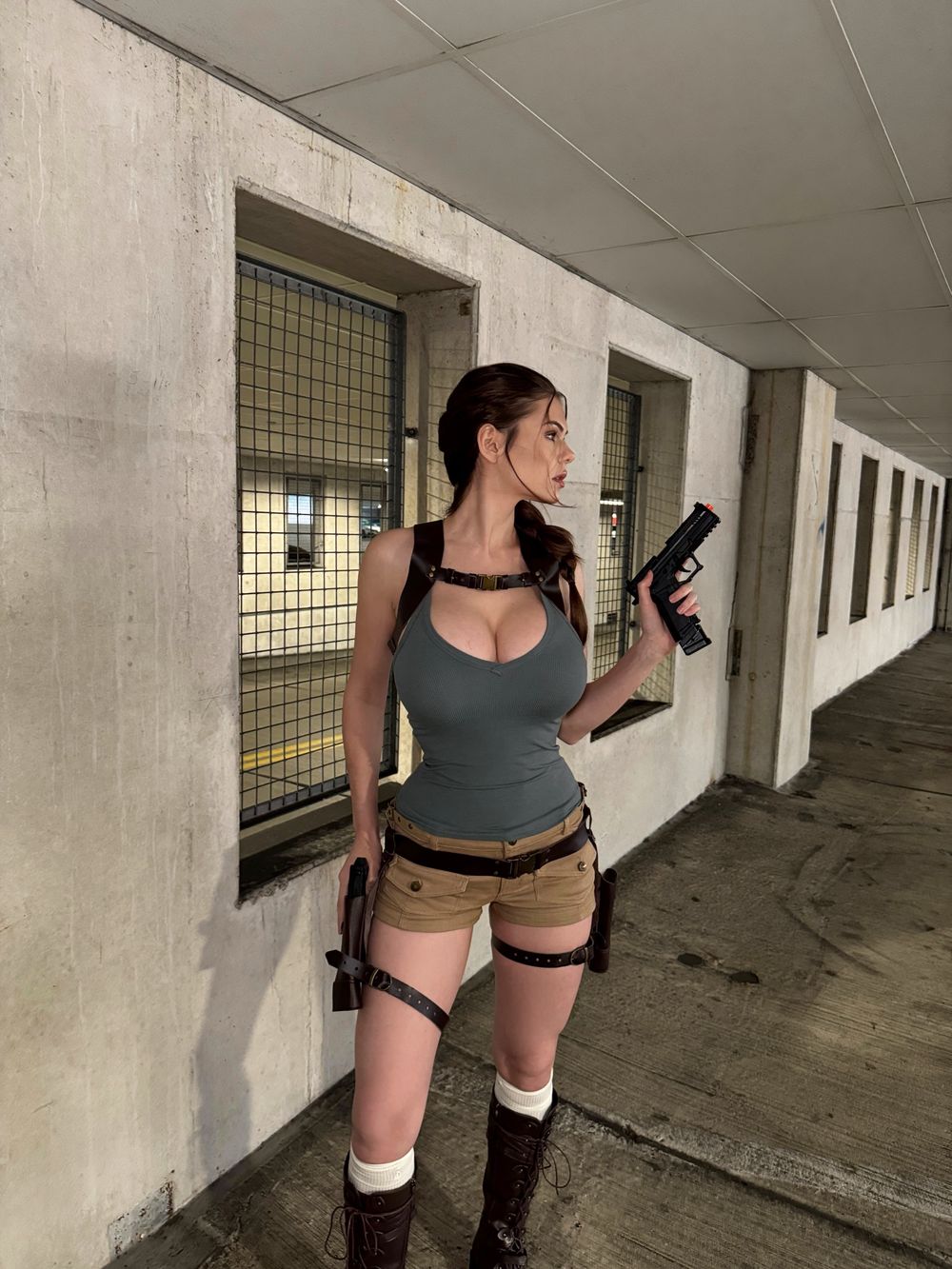 Woman with long brown hair in a braid and big breasts, dressed as Lara Croft in a tight grey tank top, khaki shorts, brown harness and holsters, stands in a concrete corridor looking to the side while holding a black prop pistol.
