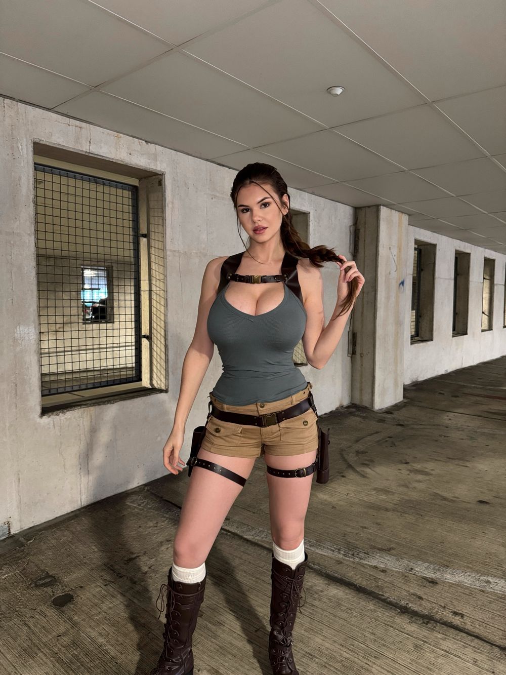 Woman with long brown hair in a braid and big breasts, dressed as Lara Croft in a tight grey tank top, khaki shorts, brown harness and holsters, stands in a concrete corridor facing the camera while holding her braid.