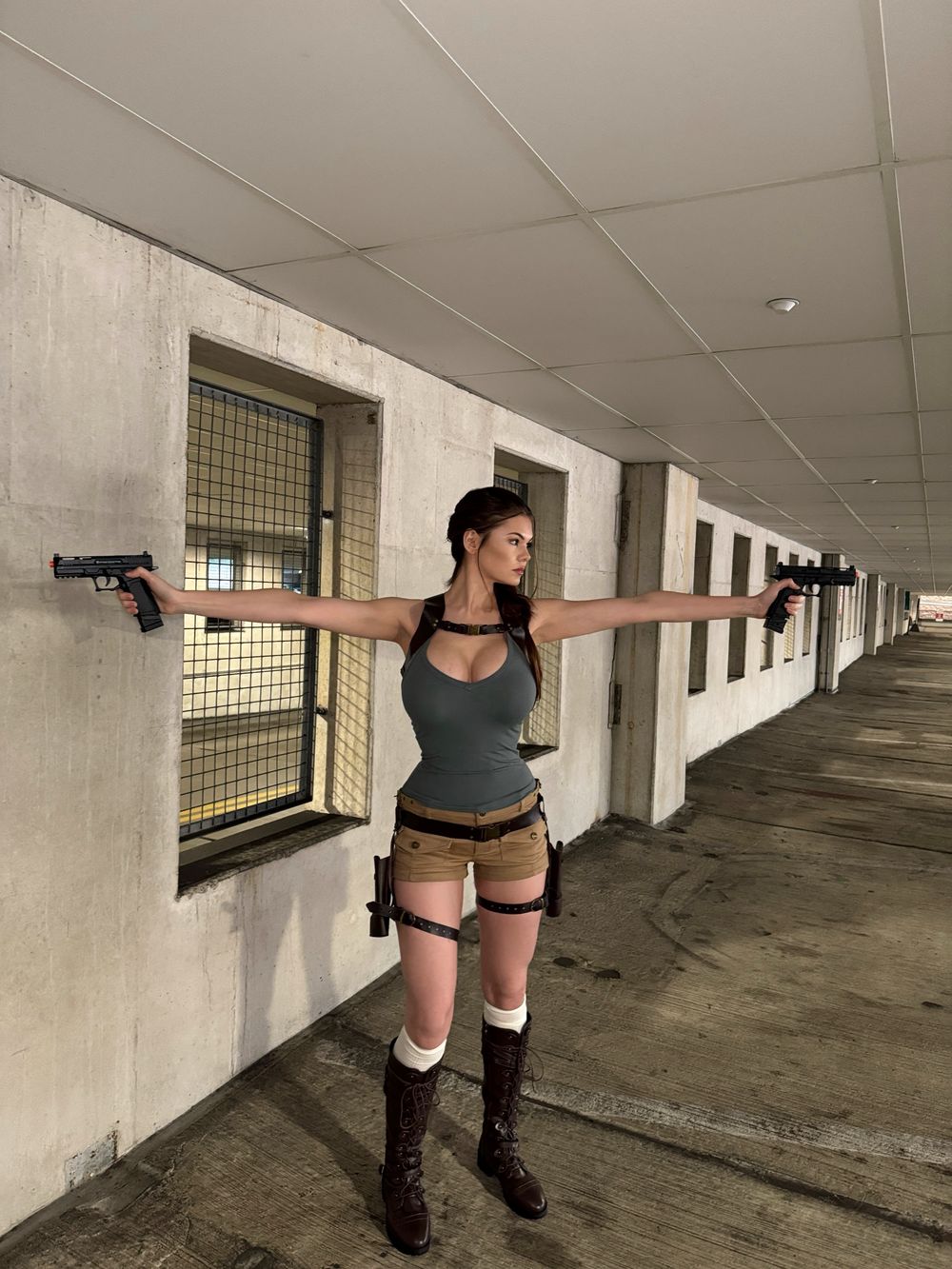 Woman with long brown hair in a braid and big breasts, dressed as Lara Croft in a tight grey tank top, khaki shorts, brown harness and holsters, stands in a concrete corridor with arms extended holding two black prop pistols.
