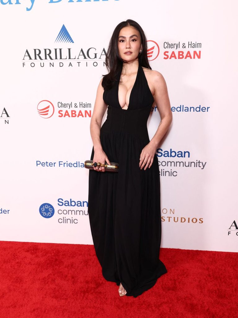 Asian woman with long dark hair wearing a black plunging halter-neck gown stands on a red carpet holding a clutch, facing the camera.