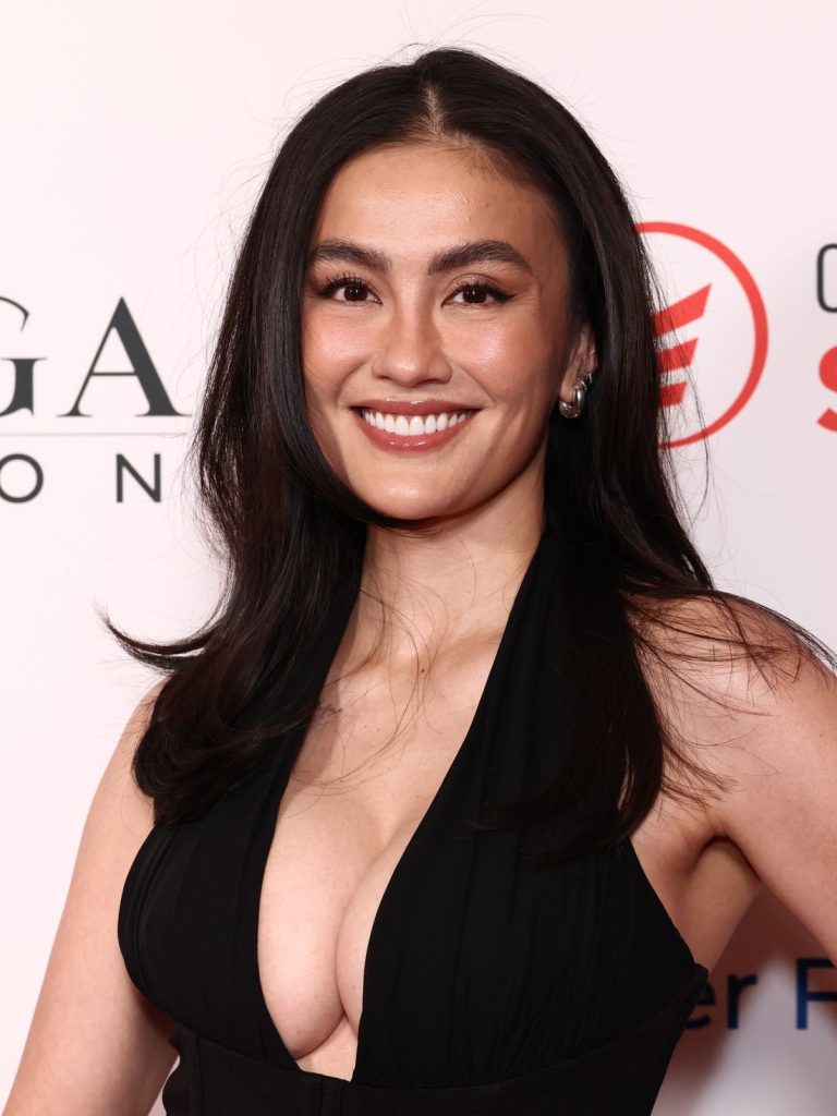 Close-up of a smiling Asian woman with full breasts wearing a black plunging halter-neck gown.