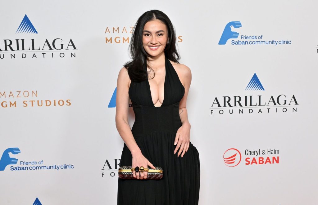 Asian woman with full breasts wearing a black plunging halter-neck gown smiles while holding a clutch in front of event logos.