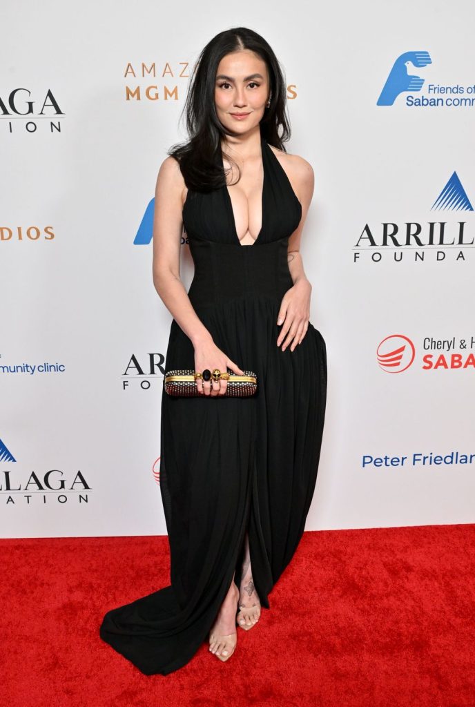 Asian woman and full breasts wearing a black plunging halter-neck gown stands on a red carpet holding a clutch.