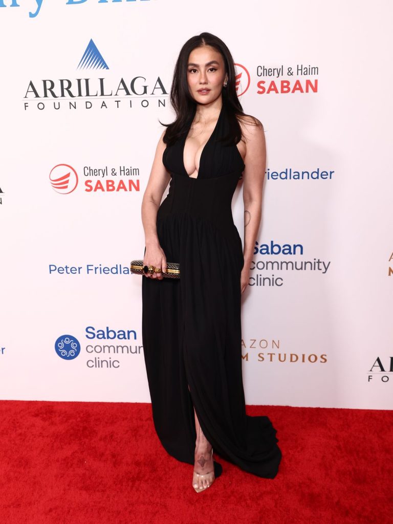 Asian woman wearing a black plunging halter-neck gown stands on a red carpet holding a clutch.