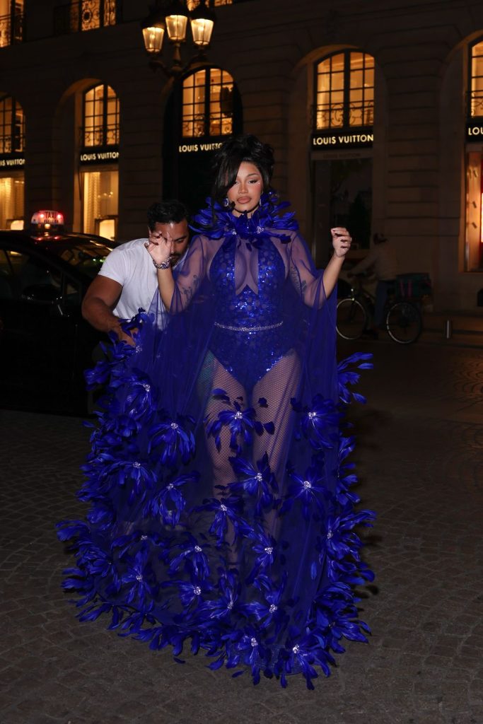 Cardi B wears a sheer, royal blue bodysuit with a matching feathered cape while stepping out of a car in front of a Louis Vuitton store.