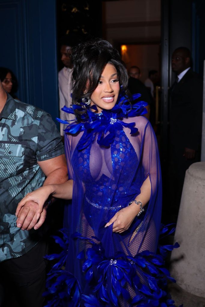 Cardi B in a sheer royal blue bodysuit with a feathered cape being escorted through a doorway.