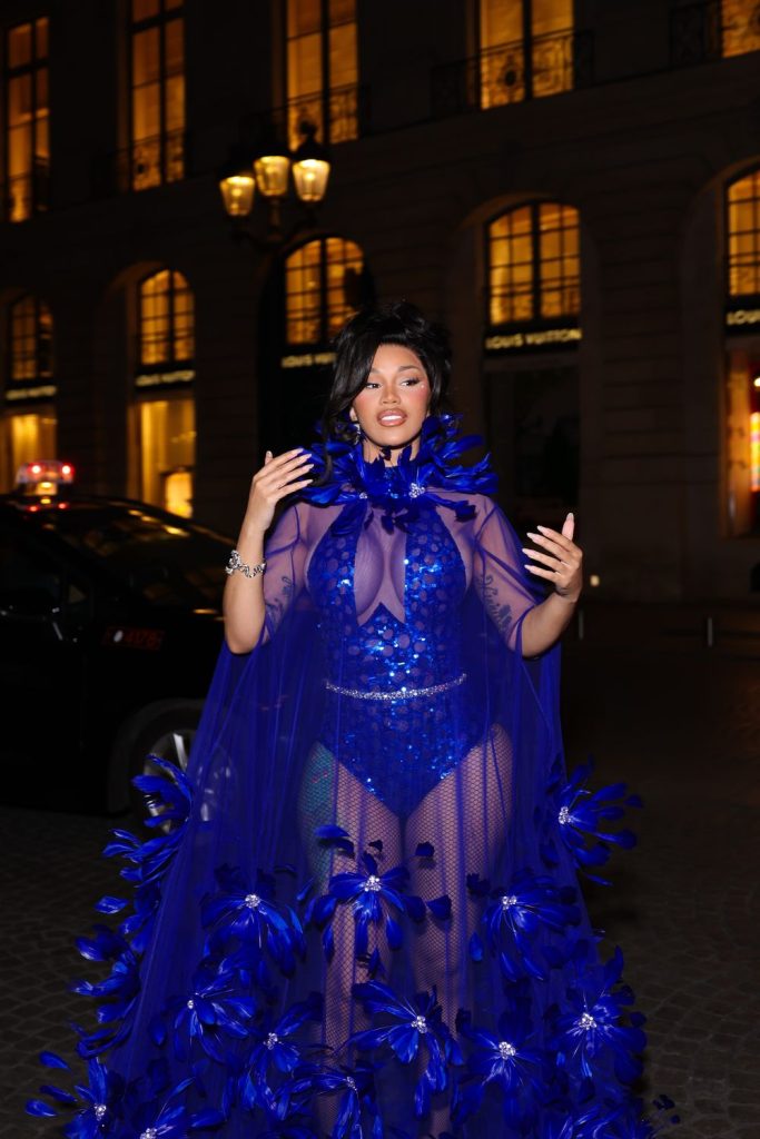 Cardi B wears a blue sheer, sequined bodysuit with a feathered cape and train while standing outside a building at night.