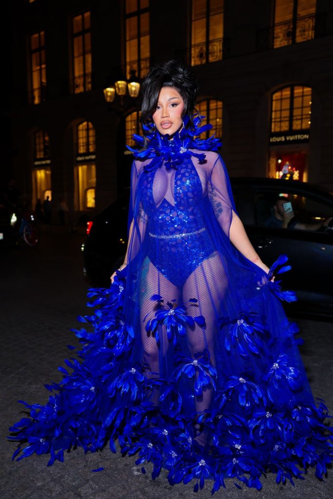 Cardi B wears a blue sheer gown with a feathered neckline and skirt, standing on a city street at night.