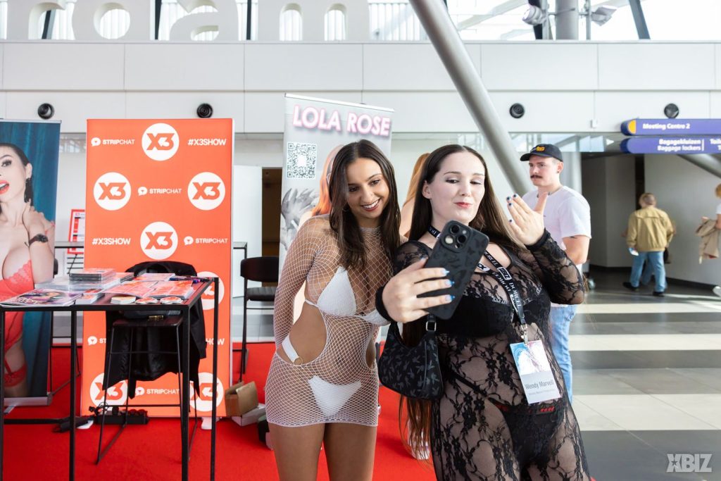 Two women in lingerie posing for a selfie at a promotional event booth with a Lola Rose sign and X3 branding in the background.