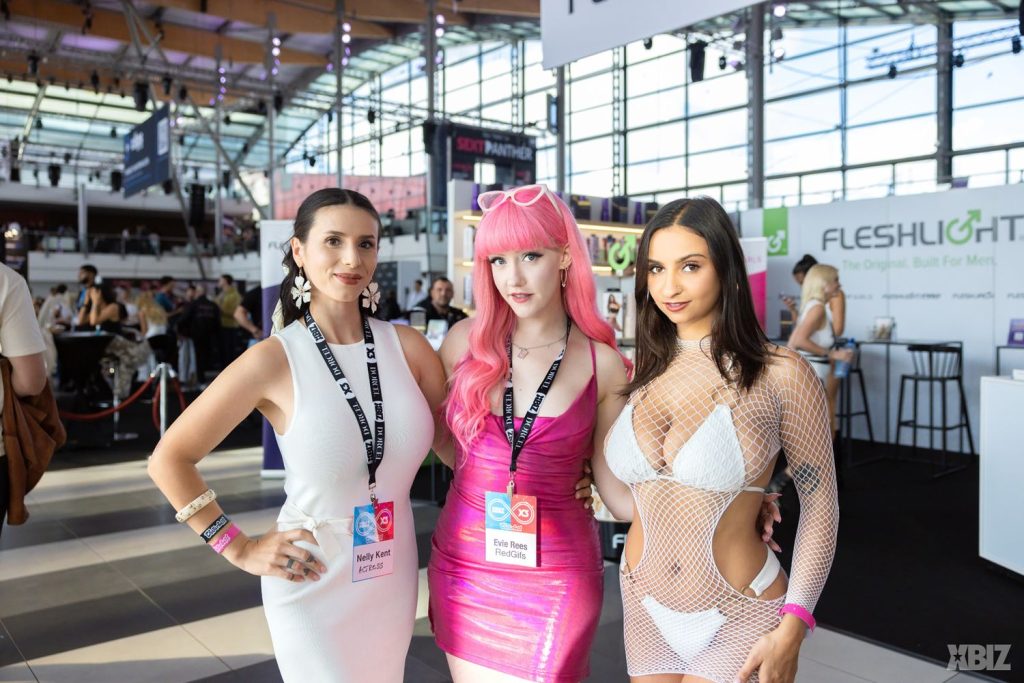 Three women posing at a convention, wearing badges labeled Nelly Kent, Evie Rees, and a third woman in a white fishnet outfit.