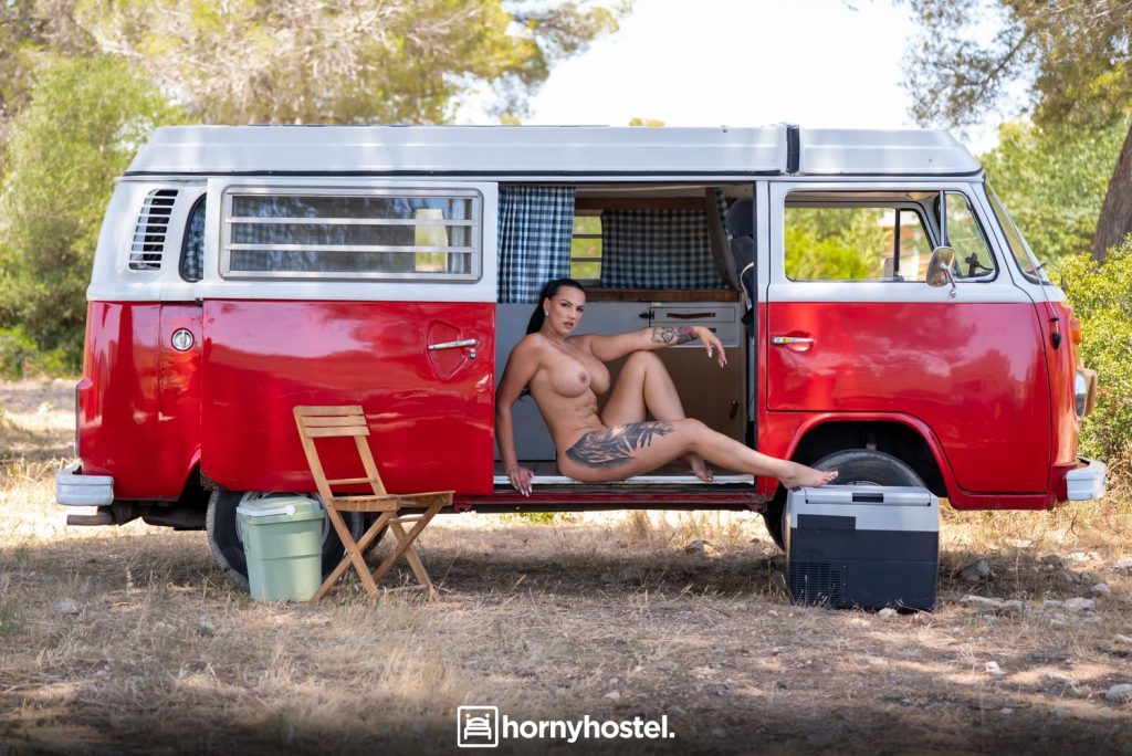 Woman with dark hair sitting nude in the open doorway of a red-and-white camper van, one leg extended, looking at the camera; outdoor setting with a wooden chair and cooler nearby.