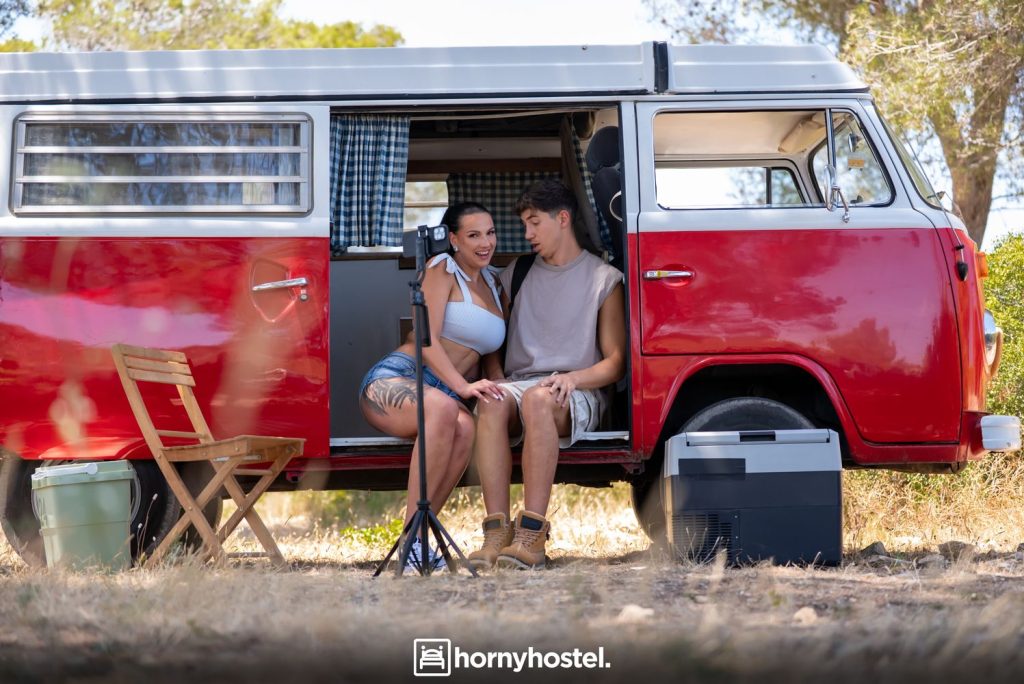 Woman with dark hair and a younger man sitting side-by-side in the open doorway of a red-and-white camper van; she wears a white striped crop top and denim shorts, he wears a sleeveless shirt and shorts, both looking at each other while a phone on a tripod records them.