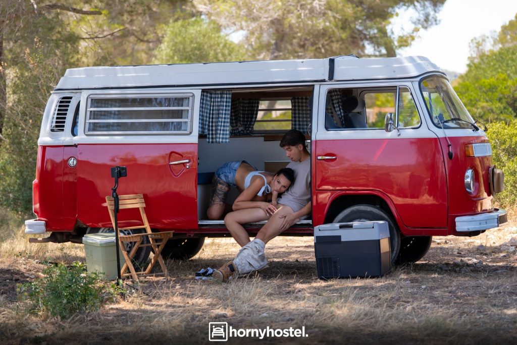 Woman with dark hair performing oral sex on a younger man seated in the open doorway of a red-and-white camper van; she wears a white striped crop top and denim shorts pulled down.