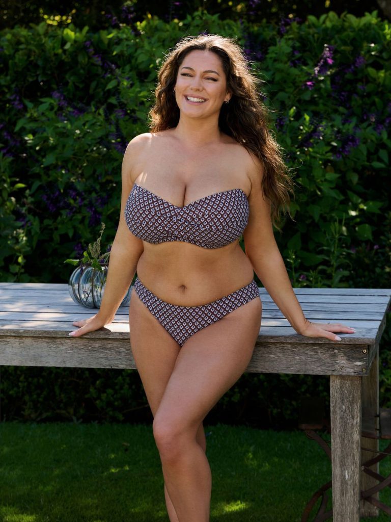 Woman with long wavy brown hair wearing a strapless brown and white geometric-print bikini, leaning against a wooden table outdoors with green foliage and purple flowers in the background.