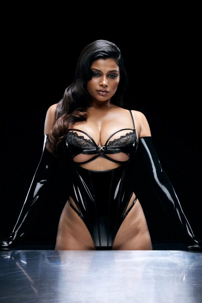 Woman with long dark wavy hair leaning forward on a metallic surface, wearing a black glossy corset with lace details and long black gloves.