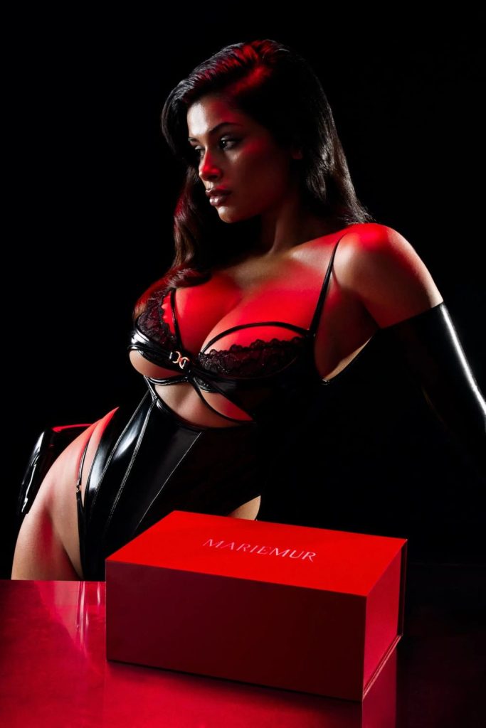 Woman with long dark hair in a black lace bra, corset, and gloves, seated beside a red box labeled MARIEMUR under red lighting.