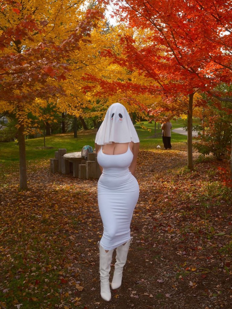 Woman wearing a white ghost-sheet mask with black eye holes and pink cheeks, a tight white sleeveless dress, and white knee-high boots, standing on a leaf-covered path in an autumn park with orange and yellow trees.