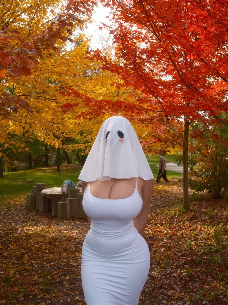 Woman wearing a white ghost-sheet mask with black eye holes and pink cheeks, a tight white sleeveless dress, and white knee-high boots, standing with hands behind her back on a leaf-covered path in an autumn park with orange and yellow trees.