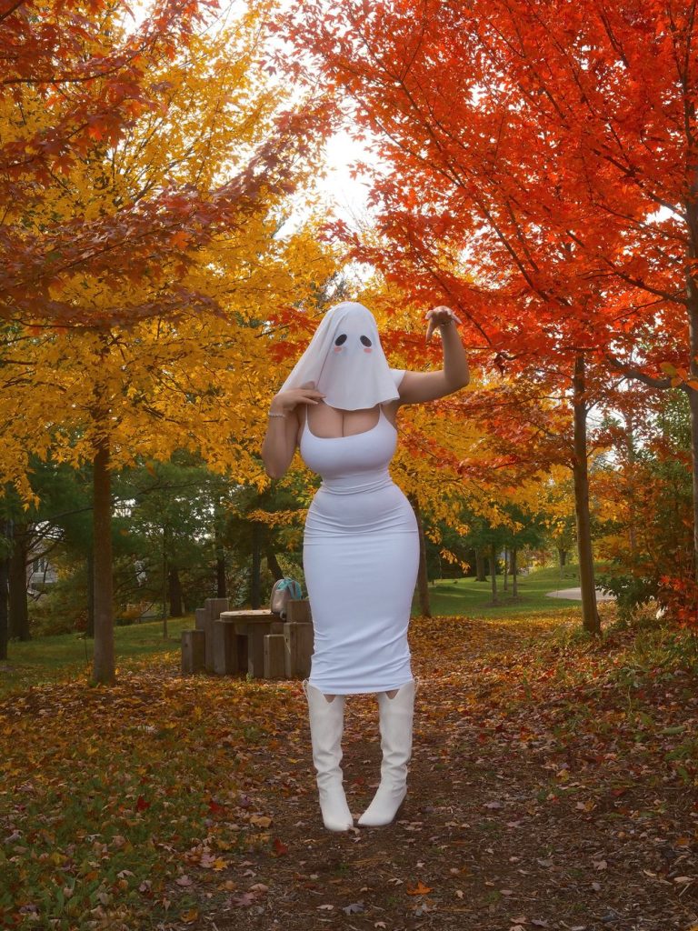 Woman wearing a white ghost-sheet mask with black eye holes and pink cheeks, a tight white sleeveless dress, and white knee-high boots, standing on a leaf-covered path in an autumn park while adjusting the mask with one hand and raising the other.