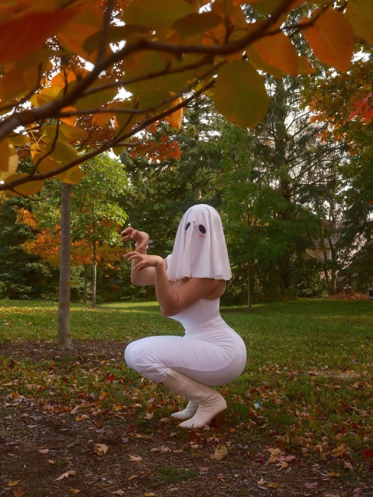 Woman wearing a white ghost-sheet mask with black eye holes and pink cheeks, a tight white sleeveless outfit, and white heeled boots, squatting on a leaf-covered ground in a park with autumn foliage, hands raised near her face.