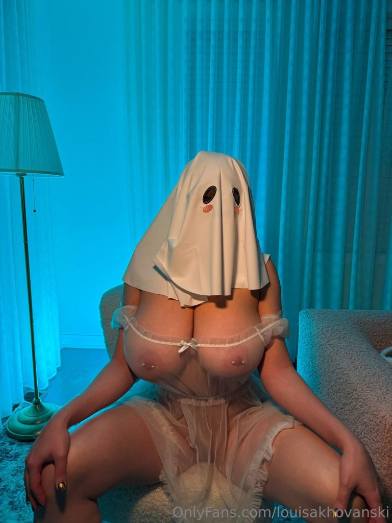 Woman seated indoors under blue lighting, wearing a white ghost-sheet mask with black eye holes and pink cheeks over a sheer white lingerie top that exposes her breasts with nipple piercings, and a matching sheer skirt.