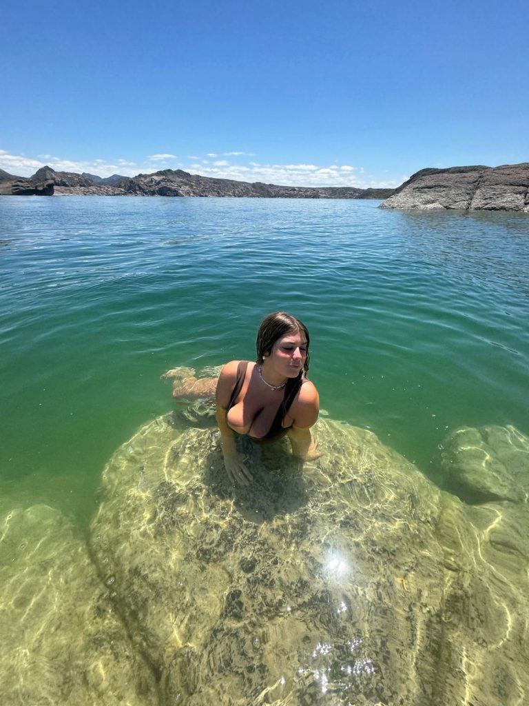 Woman with long light brown hair standing in shallow clear water in a brown bikini, looking downward.
