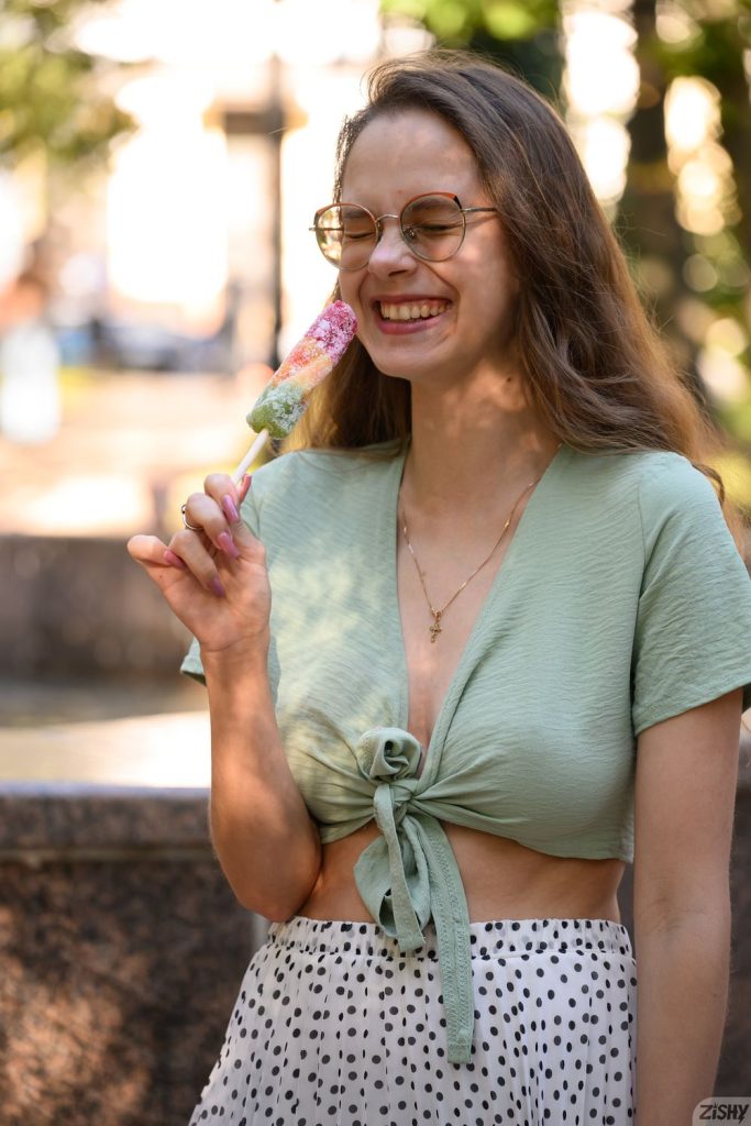 Woman with long light brown hair and glasses, wearing a light green tied crop top and white polka-dot skirt, smiling while holding a multicolored ice cream bar outdoors near a fountain.